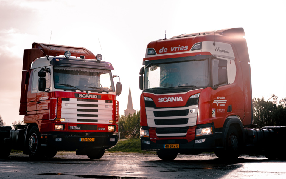 Twee rode Scania trucks van transportbedrijf de vries in Hallum in de stralende zon met op de achtergrond de torenspits van Hallum.