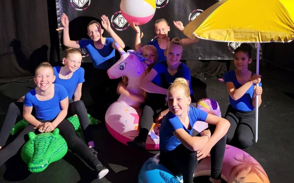 Jazz-1 dansers van Turnlust Hallum poseren vrolijk op het podium met opblaasfiguren (eenhoorn, krokodil), strandbal en gele parasol; blauwe shirts, klaar voor de show.