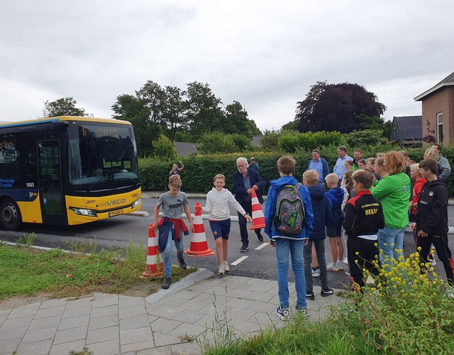 Een groep mensen, waaronder kinderen van groep 8 van basisschool It Fundamint en volwassenen, staat langs de nieuwe busbaan bij een gele Qbuzz-bus, met verkeerskegels op de weg en groen op de achtergrond.