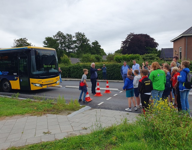 Een groep mensen, waaronder kinderen van groep 8 van basisschool it fundamint en volwassenen, luistert naar wethouder pieter braaksma die op de nieuwe busbaan spreekt met verkeerskegels voor hen geplaatst, met op de achtergrond de lijnbus te wachten op de opening.