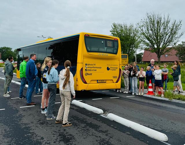 Een groep mensen, waaronder kinderen van groep 8 van basisschool It Fundamint en genodigden, staat voor een gele Qbuzz-bus op de nieuwe busbaan, met een blauwe lucht en de omgeving van Hallum op de achtergrond.