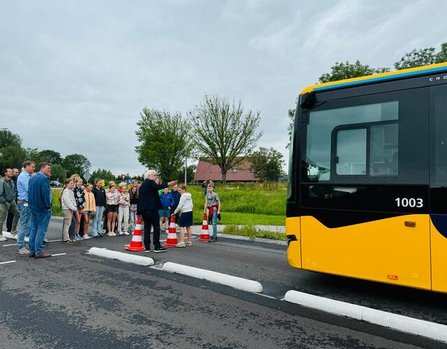 Een groep mensen, waaronder kinderen van groep 8 van basisschool It Fundamint en volwassenen, staat langs de nieuwe busbaan bij een gele Qbuzz-bus, met verkeerskegels op de weg en groen op de achtergrond.