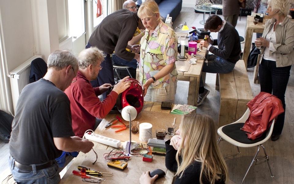 Vrijwilligers en bezoekers zitten aan tafels in gesprek tijdens een Repaircafé, met gereedschap en kapotte apparaten op tafel in een gezellige setting.