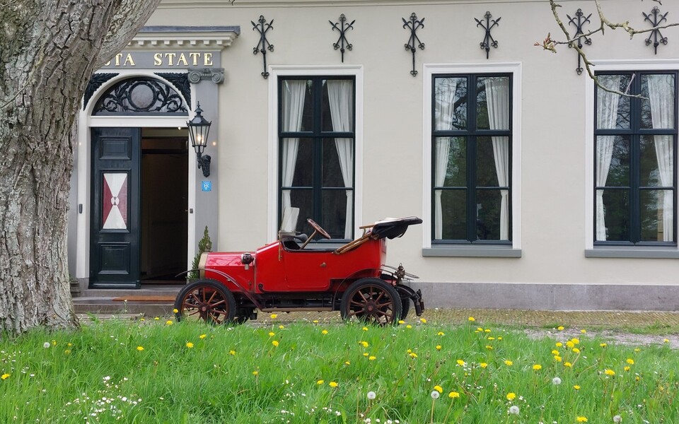 Rode oldtimerauto voor de entree van Uniastate in Hegebeintum, met gras en paardenbloemen op de voorgrond.