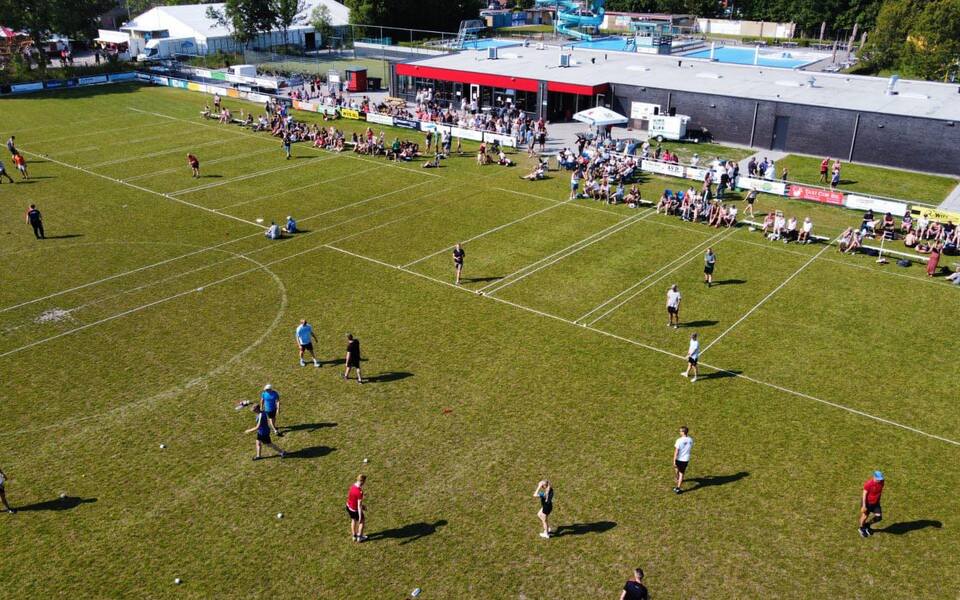 Overzichtsfoto van een kaatsveld met meerdere kaatspartijen, spelers en publiek langs de zijlijn bij het sportcomplex.