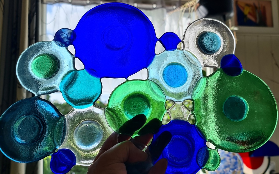 Abstract glasobject met ronde vormen in blauw, groen en transparant glas, handgemaakt fused glass kunstwerk voor het raam met lichtdoorval.