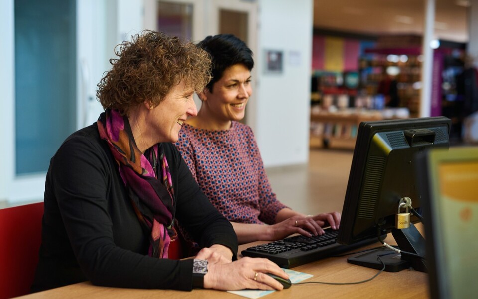 Twee vrouwen werken samen achter een computer in de bibliotheek en helpen bezoekers met digitale vragen of bibliotheekdiensten.