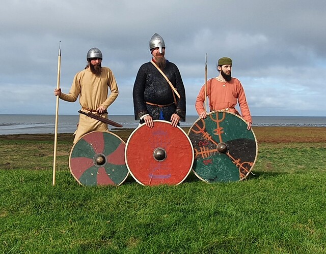 Drie vroegmiddeleeuwse strijders met speren, helmen en beschilderde schilden tijdens een reenactment.